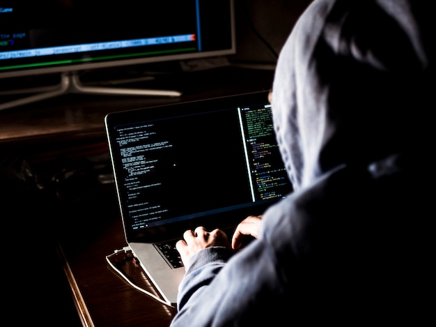 pirate informatique à capuche en tapant le code dans le programme à l'aide d'ordinateur portable dans le studio sombre