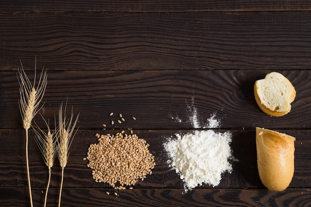 Épis de blé, céréales, farine et pain tranché sur une table en bois sombre. Vue de dessus