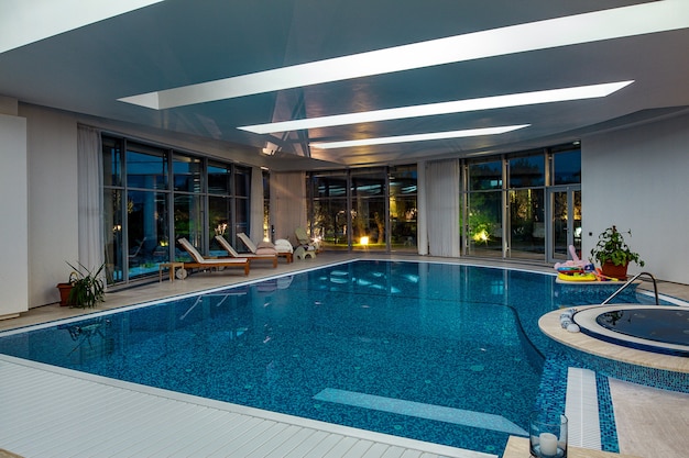 Piscine intérieure luxueuse avec baignoire jacuzzi et transats sur fond de parc verdoyant en soirée...