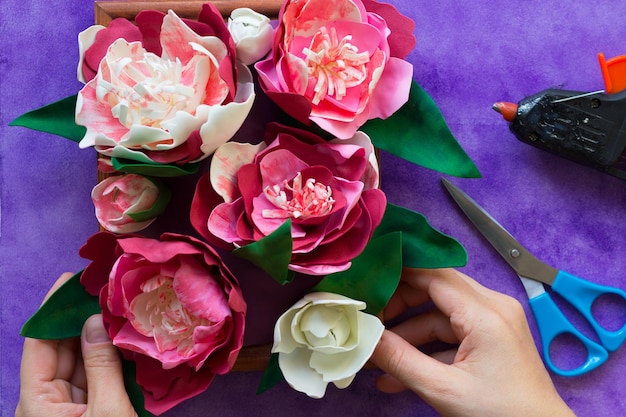 Pivoines décoratives faites à la main à base de foamiran sur panneau de bois tenant par des mains de femme et un pistolet à colle et des ciseaux