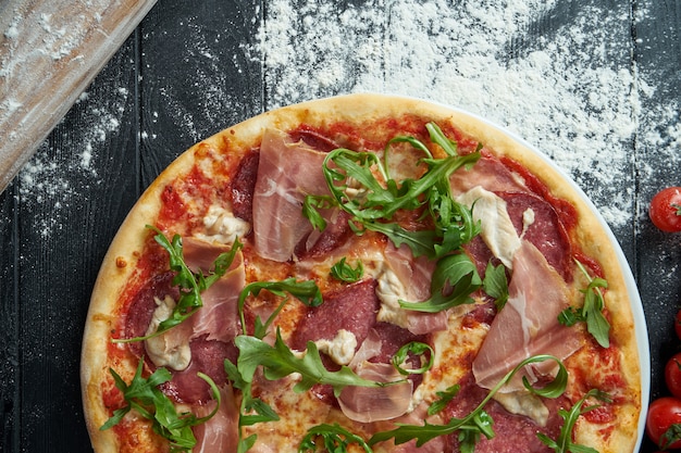 Pizza au four avec salami, prosciutto et poulet avec sauce rouge et fromage fondu sur une surface en bois noire dans une composition avec des ingrédients. Vue de dessus