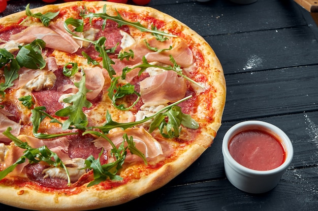 Pizza au four avec salami, prosciutto et poulet avec sauce rouge et fromage fondu sur une table en bois noire dans une composition avec des ingrédients. Vue de dessus