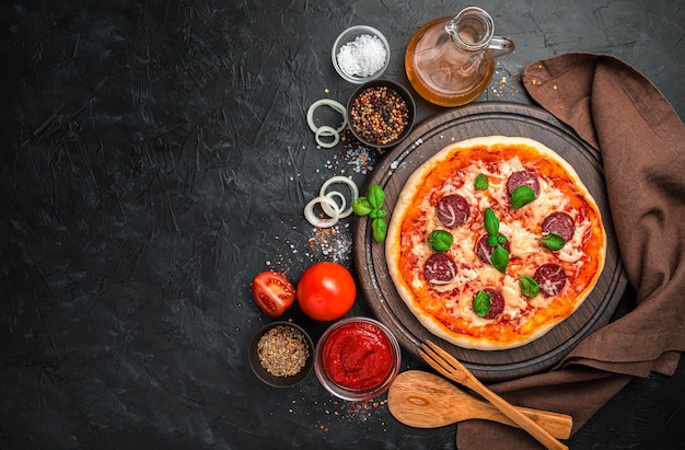 Pizza au pepperoni fraîchement préparée, tomates, sauce tomate, oignons et épices sur fond noir. Vue horizontale avec espace à copier.