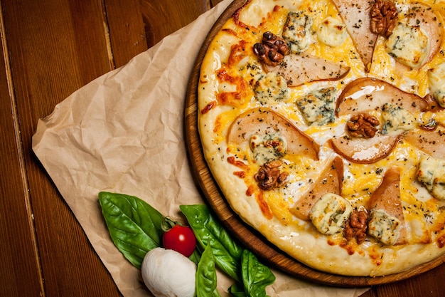 Pizza aux poires, noix, fromage Dorbl et huile de citron. Pizza maison avec poire, noix et gorgonzola sur une table en bois marron - papier kraft. A côté de la pizza se trouve un champignon, une oseille et une tomate cerise.