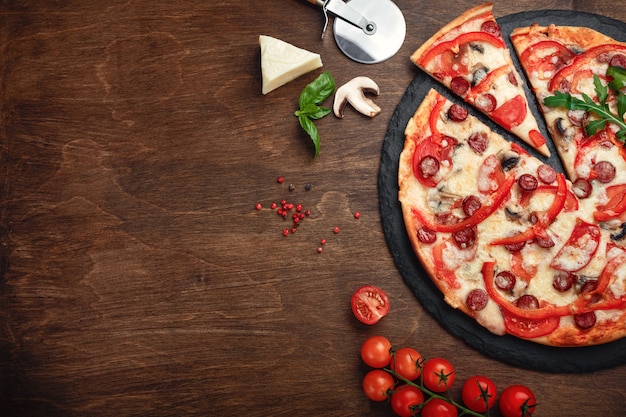 Pizza aux saucisses fumées, fromage, champignons, tomates, poivrons sur une pierre et un plateau en bois brun.