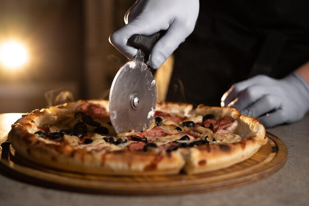 La pizza chaude au fromage est coupée à la main par un chef portant un gant en caoutchouc avec un couteau spécial. Produits de boulangerie prêts à l'emploi dans une pizzeria.