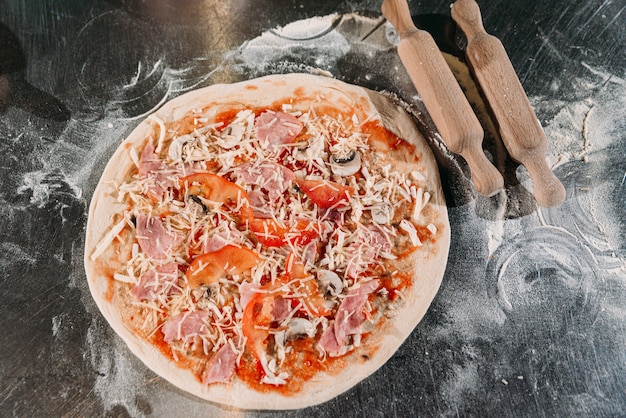 Une pizza crue farcie au fromage, aux tomates et au bacon se trouve sur la table de la cuisine, à côté des rouleaux à pâtisserie. La farine est versée sur la table. Vue de dessus, gros plan