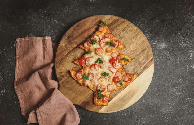 Une pizza en forme d'arbre de Noël sur une planche à découper avec une serviette de cuisine se trouve à gauche sur un fond sombre