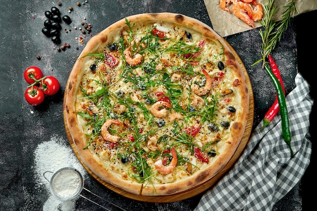 Pizza italienne au four avec sauce, fromage, roquette, crevettes et olives dans une composition avec des ingrédients sur une table sombre. Vue de dessus Pizza aux fruits de mer