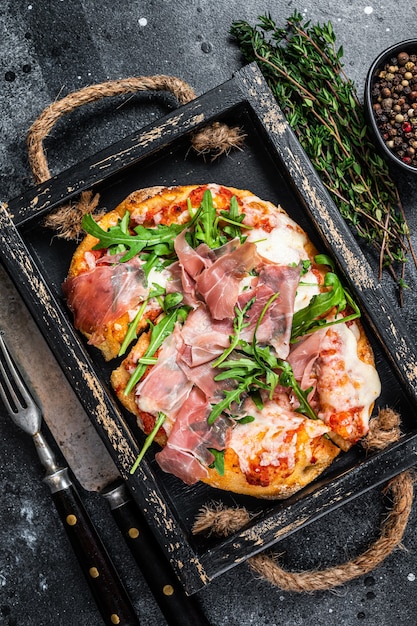 Pizza italienne au jambon de Parme prosciutto, salade de roquette et fromage dans un plateau en bois rustique. Fond noir. Vue de dessus.