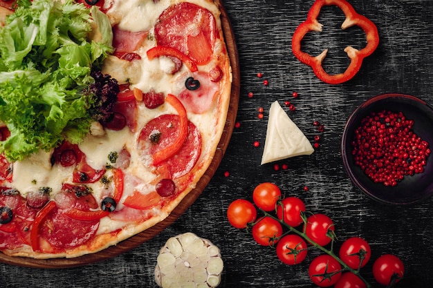 Pizza avec jambon, salami, fromage, champignons, tomates cerises, poivrons et salade sur un tableau noir.