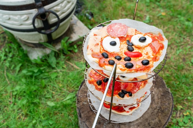 Pizza maison amusante pour enfants avec yeux au fromage visage olives noires bouche tomate sauce saucisse Cuisine familiale en plein air dans un four oriental tandoor Grille métallique spéciale pour légumes barbecue Herbe verte