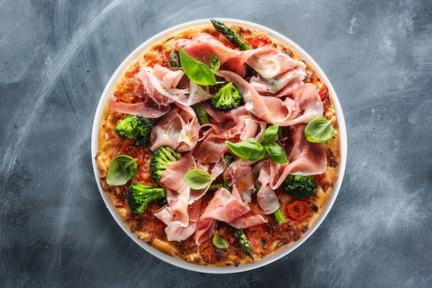 Pizza maison avec viande, brocoli et basilic sur une surface bleue.