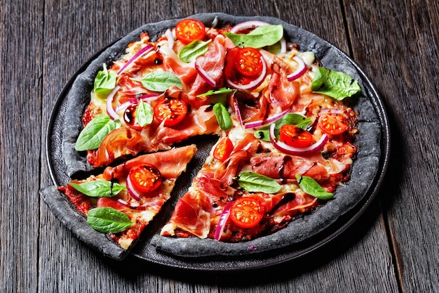 Pizza à la pâte noire avec tranches de jambon, fromage mozzarella, tomate, feuilles d'épinards et oignon rouge sur un plateau noir sur une table en bois vue horizontale d'en haut