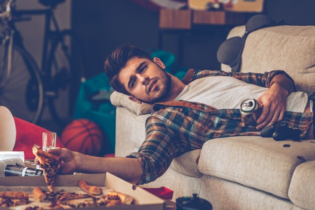 La pizza peut-elle aider avec la gueule de bois? Jeune bel homme tenant une tranche de pizza et une canette de bière