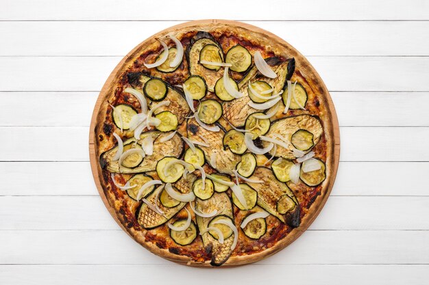 Pizza végétarienne entière garnie de courgettes aubergines grillées et d'oignons sur une plaque en bois vue de dessus sur fond de tableau blanc