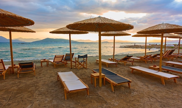 Plage avec chaises longues et parasols en paille au bord de la mer au coucher du soleil, l'île d'Egine, Grèce. Vue panoramique grand angle