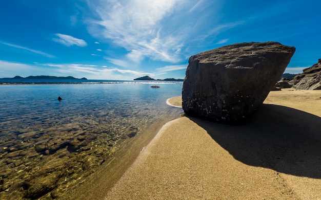 Une plage avec un gros rocher dessus et le ciel est bleu et l'eau est claire.