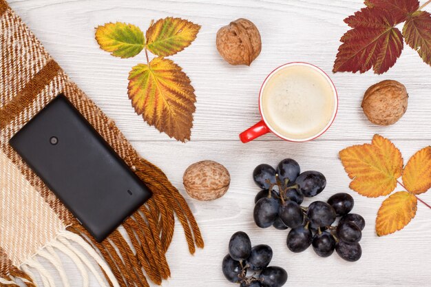 Plaid à carreaux, téléphone portable, feuilles sèches jaunes et brunes, tasse de café, raisins et noix sur fond en bois. Une composition d'automne. Vue de dessus.