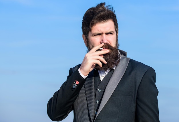 Plaisir de fumer Habitude relaxante mais nocive Homme avec barbe et moustache tenir une cigarette Hipster barbu fumant une cigarette sur fond de ciel bleu Un gars avec une cigarette profite de l'influence de la nicotine