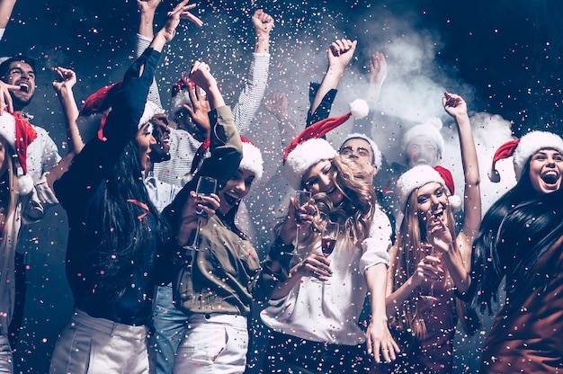 Plaisir de Noël. Groupe de beaux jeunes en chapeaux de Père Noël jetant des confettis colorés et ayant l'air heureux