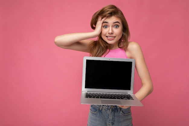 Plan de la charmante jeune femme surprise tenant un ordinateur portable avec un écran vide portant un t-shirt rose regardant la caméra