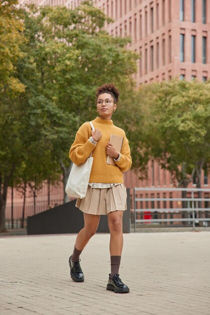 Plan complet d'une étudiante séduisante en jupe-pull tricoté jaune et chaussures portant un sac en tissu contenant des manuels