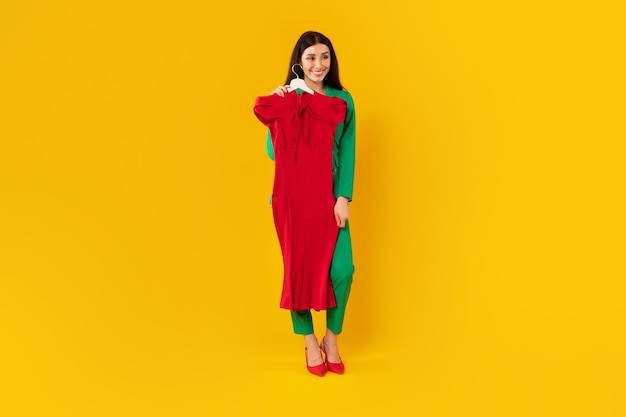 Plan complet d'une femme de créateur de mode debout avec un cintre à la main sur une robe élégante rouge sur fond jaune