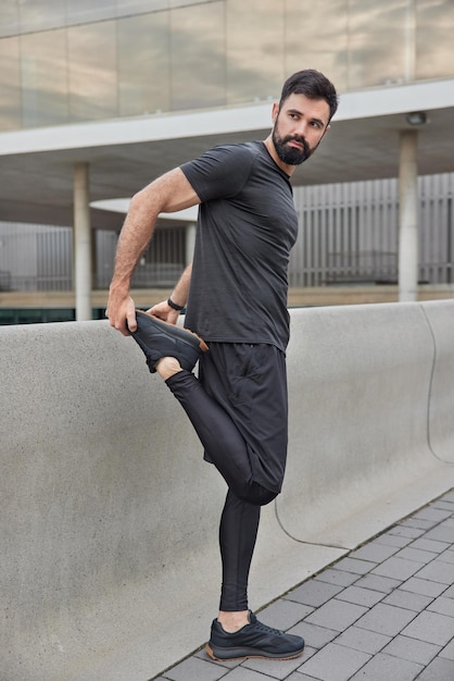 Plan complet d'un homme barbu sportif adulte étire les jambes avant de courir se réchauffe se prépare pour l'entraînement vêtu de vêtements de sport regarde ailleurs pose à l'extérieur a des bras musclés bonne condition physique