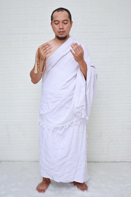 Plan complet d'un homme musulman asiatique portant des vêtements blancs d'ihram et priant tout en tenant la prière