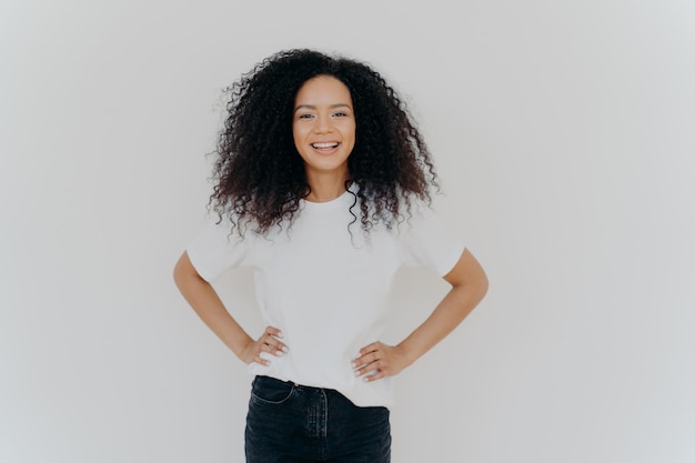 Plan demi-longueur d'une belle fille gaie du millénaire se tient avec une expression de visage heureux sur fond de studio blanc garde les deux mains sur la taille a les cheveux bouclés maquillage minimal silhouette mince