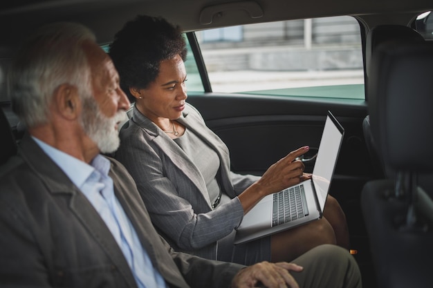Plan de deux personnes multiethniques prospères utilisant un ordinateur portable et discutant assis à l'arrière d'une voiture pendant leur trajet professionnel.