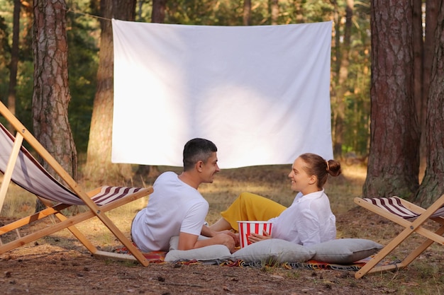 Plan extérieur d'un couple allongé dans la forêt avec rétroprojecteur avec écran blanc vierge mangeant du pop-corn parlant et profitant de leur temps libre ensemble dans la nature