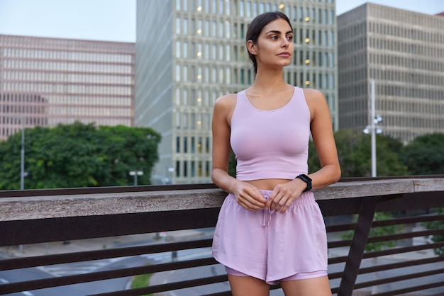 Plan extérieur d'une femme sportive vêtue de vêtements de sport concentrée avec une expression réfléchie pose sur un pont contre des gratte-ciel pendant la journée fait régulièrement du sport Mode de vie sain