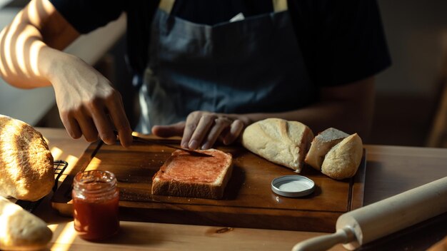 Plan d'un homme portant un tablier répandant de la confiture de framboises sur une tranche de pain avec un couteau de table sur une table de cuisine en bois