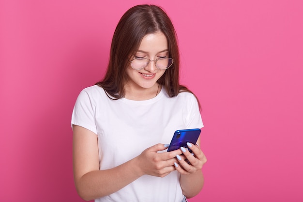 Plan horizontal d'adolescent adorable aux cheveux longs, tenant un téléphone portable moderne, fait défiler les réseaux sociaux, a une expression joyeuse, porte des lunettes et un t-shirt décontracté, isolé sur rose