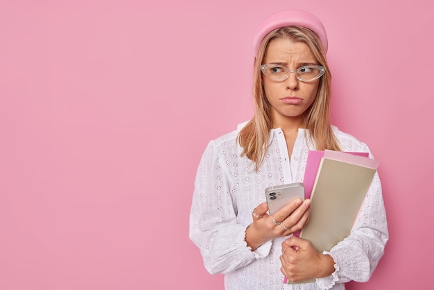 Plan horizontal d'une adolescente contrariée triste d'avoir des problèmes d'étude se prépare aux examens universitaires tient un téléphone portable avec une expression sombre porte un chemisier blanc et des lunettes pose à l'intérieur