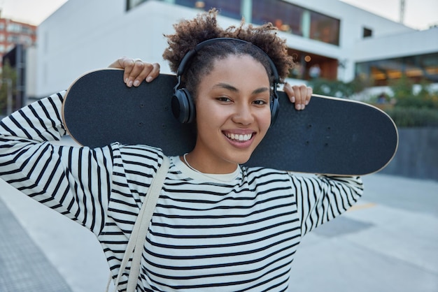 Plan horizontal d'une adolescente heureuse avec deux chignons tenant une planche à roulettes derrière vêtue d'un pull rayé décontracté écoute la piste audio dans les écouteurs recrée en ville