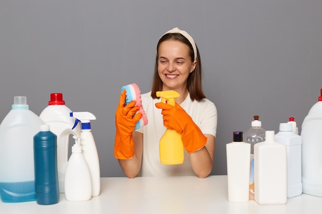 Plan horizontal d'une adorable femme heureuse souriante portant un t-shirt blanc et des gants orange posant isolé sur fond gris pulvérise du détergent sur l'éponge démontre l'utilisation du nettoyant