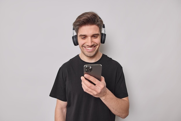 Plan horizontal d'un beau jeune homme heureux télécharge des chansons audio sur une liste de lecture porte des écouteurs sans fil sur les oreilles sourit joyeusement vêtu d'un t-shirt noir décontracté isolé sur fond blanc