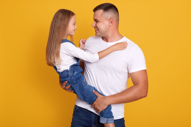 Plan horizontal d'un beau père portant sa petite fille, des modèles posant isolés sur un mur jaune, papa et son enfant se regardant, habillés de vêtements décontractés. Concept de famille.