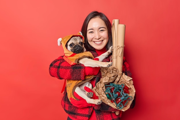 Plan horizontal d'une belle femme asiatique aux cheveux noirs et joyeuse embrasse un chien carlin vêtu de vêtements d'hiver avec amour porte des décorations pour les vacances a la bonne humeur isolée sur fond rouge vif