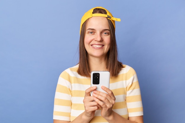Plan horizontal d'une blogueuse adolescente satisfaite portant un t-shirt rayé et une casquette de baseball debout isolé sur fond bleu à l'aide d'un téléphone portable regardant la caméra avec un sourire à pleines dents