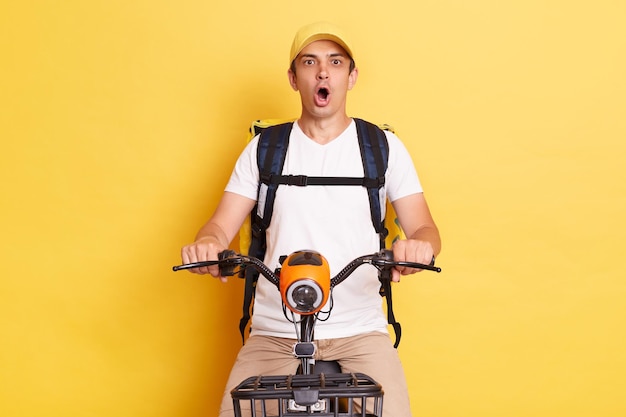 Plan horizontal d'un coursier surpris choqué faisant du vélo avec un sac à dos livrant un repas à partir d'un café ou d'un restaurant à emporter regardant la caméra avec la bouche ouverte isolée sur fond jaune