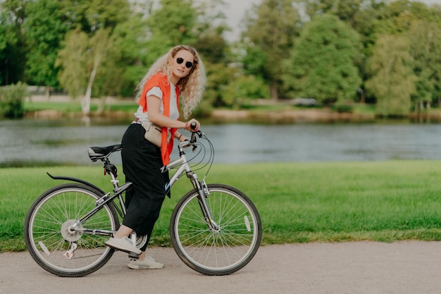 Plan horizontal d'une cycliste féminine active porte un t-shirt pantalon noir et des baskets s'arrête près du bord de la rivière garde les pieds sur les pédales du vélo belle nature pittoresque en arrière-plan Journée ensoleillée repos actif