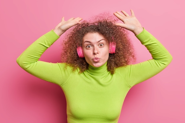 Photo plan horizontal d'une femme aux cheveux bouclés garde les lèvres pliées lève les bras porte des écouteurs sans fil col roulé vert isolé sur un mur rose écoute une nouvelle piste apprécie la musique cool de la liste de lecture