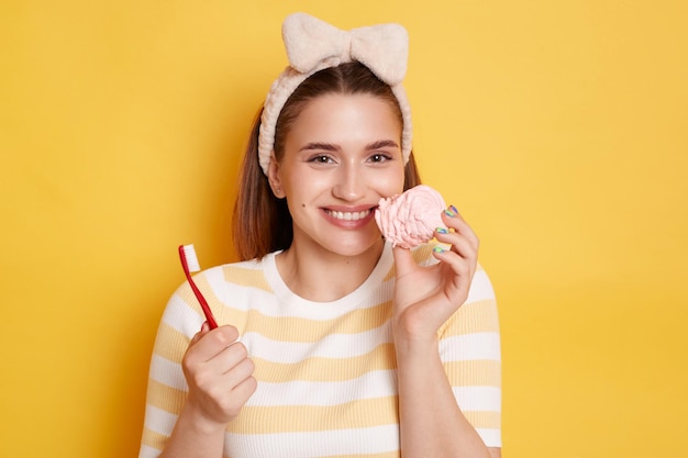 Plan horizontal d'une femme aux cheveux noirs souriante et satisfaite portant une chemise rayée et une bande de cheveux posant isolé sur fond jaune tenant une brosse à dents et une guimauve savoureuse n'a pas de problèmes avec les dents