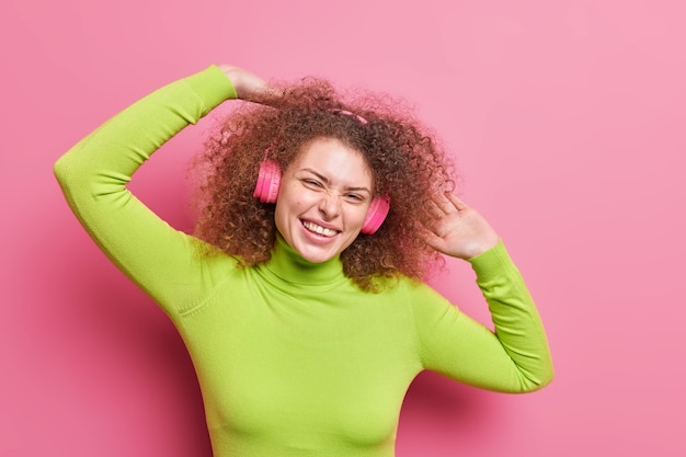 Plan horizontal d'une femme européenne positive aux cheveux bouclés lève les bras incline la tête vêtue d'un col roulé vert décontracté écoute la piste audio via des écouteurs isolés sur un mur rose. Mode de vie