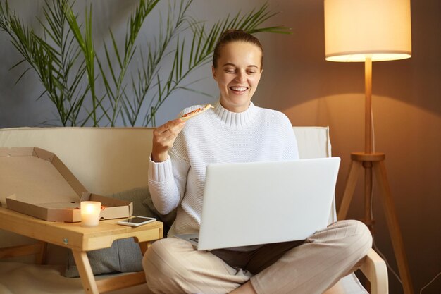 Plan horizontal d'une femme joyeuse et affamée avec une coiffure chignon portant un chandail blanc assis sur la toux et travaillant sur un ordinateur portable travaillant en ligne et prenant une collation en mangeant de la pizza