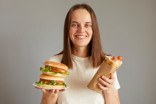 Plan horizontal d'une femme mangeant de la restauration rapide tenant un hot-dog et un hamburger portant un t-shirt blanc posant isolé sur fond gris aime tellement les aliments malsains